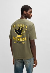 Oliven grøn bomulds t-shirt med et sort grafisk design med flammer, "CALIFORNIA," og "HOTHEADZ" i gult på ryggen. Korte ærmer, rund hals.