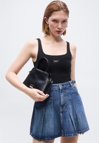 Jeune femme portant un débardeur noir et une jupe plissée en denim, tenant un petit sac à main noir devant un fond uni.