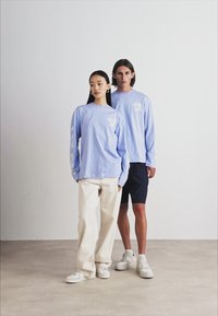 Long-sleeve light blue t-shirts with printed accents, paired with cream baggy pants and dark shorts. Both models wear white sneakers.