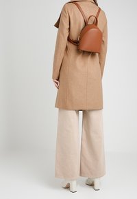 Mochila de cuero marrón con parte superior redondeada, llevada en la espalda. Combinada con un abrigo beige y pantalones de pierna ancha. Diseño minimalista, sin herrajes visibles.