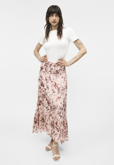 Kvinde med langt mørkt hår iført hvid kortærmet top, lyserød blomstret maxinederdel og hvide sandaler med stropper, stående med hænderne på hofterne.