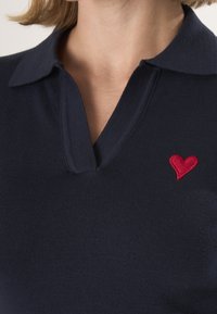 Marineblauwe poloshirt met een kraag en korte mouwen, voorzien van een kleine rode hartjesborduring aan de linkerkant. Zachte stofstructuur.