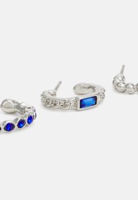 Três brincos de prata: um apresenta uma pedra retangular azul, um tem adornos azuis e um tem um design liso e redondo.