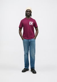 Bordové bavlnené tričko s logom "Calvin Klein", skombinované so svetlomodrými džínsami a čiernymi topánkami. Modelka má na hlave krémovú pletenú čiapku.