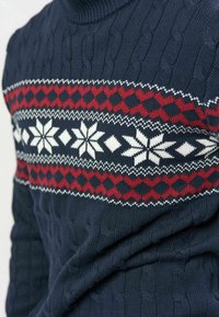 Pull en maille bleu marine avec motif losange rouge et flocon de neige blanc sur la poitrine et texture en maille torsadée.