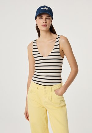 Frau mit blauem Cap, ärmellosem weißen Top mit schwarzen horizontalen Streifen und hoch taillierter gelber Hose, steht mit einer Hand in der Tasche.