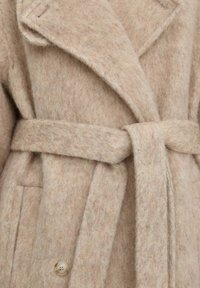Béžový vlněný kabát se širokými klopami, zavazovacím pásem v pase a viditelným knoflíkem blízko spodní části na přední straně.