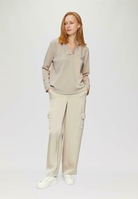 Beige Bluse mit langen Ärmeln und V-Ausschnitt, kombiniert mit beige Cargo-Hosen mit Seitentaschen und weißen Sneakers mit strukturierten Sohlen.