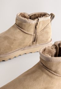Bottes chevilles en daim beige avec fermeture éclair, doublure moelleuse et semelle en caoutchouc texturée. Présentent un détail de couture subtile et une languette de tirage.