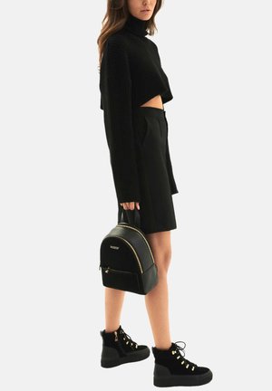 Schwarzer, cropped Rollkragenpullover mit geripptem Textur, kombiniert mit einem schwarzen Rock. Ergänzt durch einen schwarzen Rucksack und schwarze Schnürstiefel.