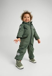 Groene waterdichte jumpsuit met een rits, zijzakken en reflecterende accenten; textuurstof, met capuchonontwerp, gecombineerd met groene schoenen.