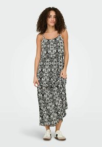 Femme aux cheveux bouclés portant une robe longue sans manches à motifs noir et blanc et des baskets blanches, debout devant un fond uni.