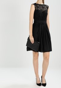 Robe noire avec un corsage en dentelle, design sans manches, taille cintrée et jupe évasée. Associée à une pochette noire et des talons pointus.