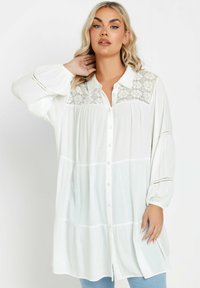 Blusa bianca con bottoni, con dettagli in pizzo sullo scollo, pieghe sotto, maniche ampie e leggere variazioni di texture nel tessuto.