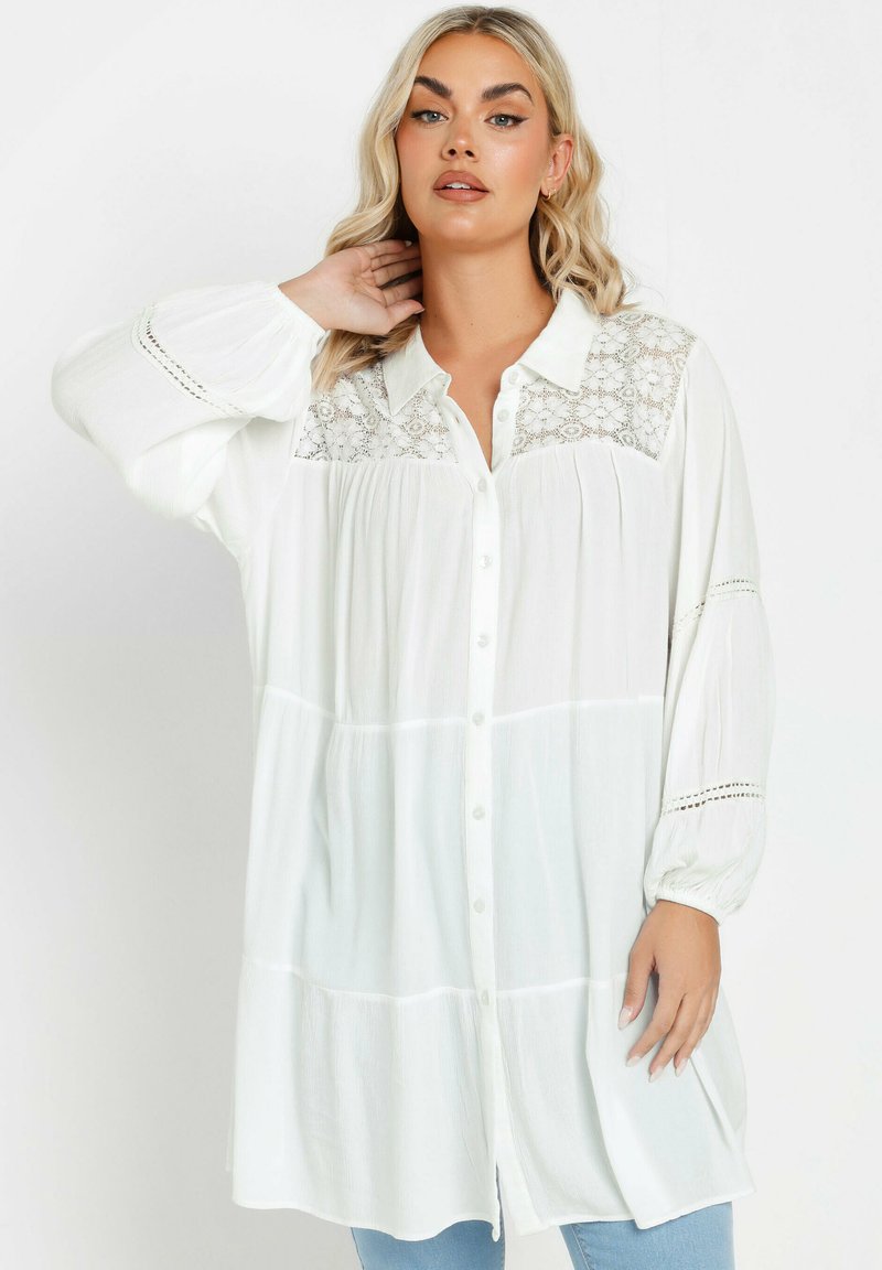 Blusa bianca con bottoni, con dettagli in pizzo sullo scollo, pieghe sotto, maniche ampie e leggere variazioni di texture nel tessuto.