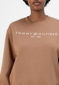 Hnědá mikina se žebrovaným kulatým výstřihem, s bílým logem "TOMMY HILFIGER" a textem "EST. 1985". Měkká tkanina s jemnou strukturou.
