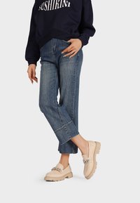 Mocassini beige senza lacci con superficie testurizzata, caratterizzati da un accento decorativo a catena, abbinati a jeans blu a vita alta e una felpa scura.