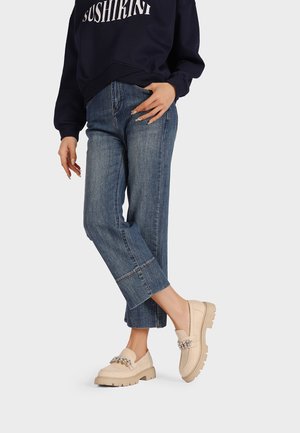 Mocassini beige senza lacci con superficie testurizzata, caratterizzati da un accento decorativo a catena, abbinati a jeans blu a vita alta e una felpa scura.