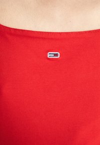 Czerwona bawełniana bluzka na ramiączkach z kwadratowym dekoltem i prążkowaną teksturą. Posiada małe, haftowane logo na piersi w kolorze niebieskim, białym i czerwonym.