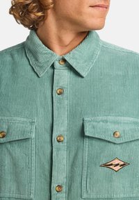 Mint green corduroy shirt featuring a pointed collar, two chest pockets, wooden buttons, and a diamond-shaped embroidered patch.