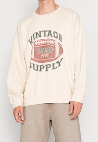 Osoba oblečená v krémovém svetru s vintage fotbalovou grafikou a textem "VINTAGE SUPPLY 1991", v kombinaci se světlými béžovými kalhotami.