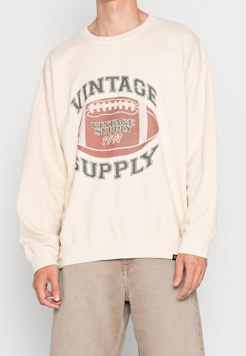 Osoba oblečená v krémovém svetru s vintage fotbalovou grafikou a textem "VINTAGE SUPPLY 1991", v kombinaci se světlými béžovými kalhotami.