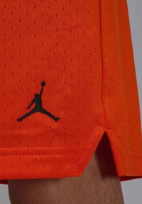 Lyse oransje basketballshorts laget av pustende perforert stoff, med en svart Jumpman-logo nær kanten og sideåpninger.