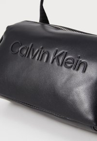 Černá kabelka z umělé kůže s vyraženým logem "Calvin Klein", hladkým povrchem a stříbrným zipem nahoře. Kompaktní obdélníkový tvar.