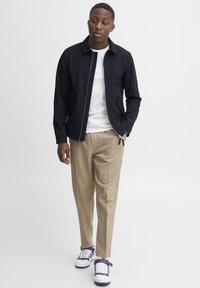 Giacca navy con zip sopra una maglietta bianca, pantaloni beige a trama e sneakers bianche con dettagli navy. Design a gamba dritta, stile minimalista.