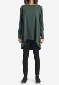 Tunic verde a maniche lunghe in tessuto morbido, con scollo rotondo e orlo asimmetrico, abbinata a pantaloni neri attillati e scarpe slip-on nere.