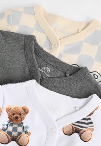Drei Babykleidungsteile: ein beiges kariertes Hemd, ein graues Hemd und ein weißes Hemd mit einem Teddybär-Graphic. Baumwollstoff, runder Ausschnitt.