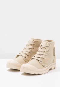 Beige vysoké tenisky vyrobené z plátna s gumovou špičkou a strukturovanou podrážkou, s tkaničkami a kruhovým logem na boku.