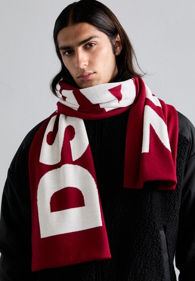 Écharpe rouge avec des lettres blanches en gras. Fabriquée en tissu tricoté, elle a une texture douce et est enroulée autour du cou sur une veste en polaire noire.
