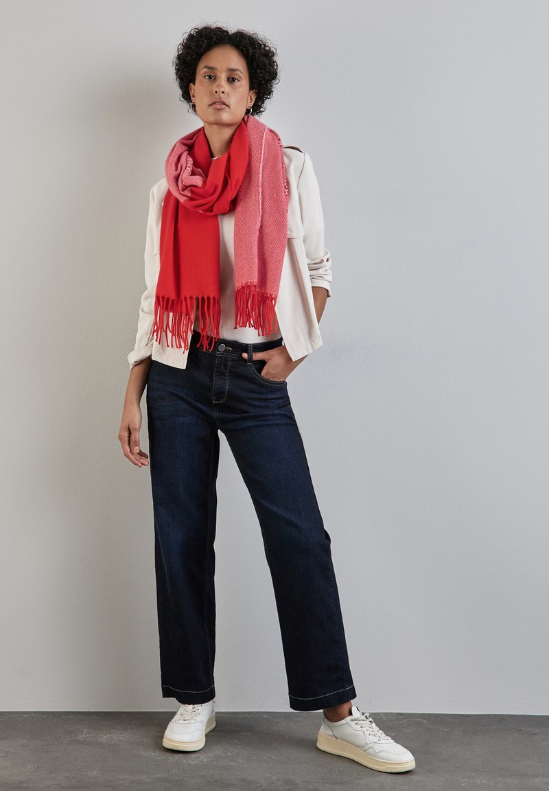 Rode en roze wolmix sjaal met franjes, witte lange mouwen shirt, donkere denim wijde jeans en witte sneakers.