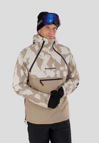 Beige-weiße Camouflage-Skijacke mit einer Frontreißverschluss-Tasche, hohem Kragen und schwarzen Akzenten. Das Model trägt schwarze Handschuhe und eine Skibrille.