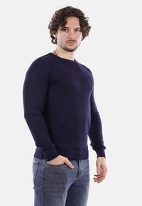 Maglione lavorato a maglia blu navy con scollatura rotonda e polsini a coste, design aderente, indossato con jeans azzurri. Motivo testurizzato sul maglione.