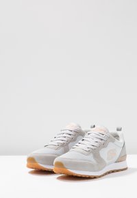 Ett par gråa och vita sneakers med gummisulor, nät- och mockadetaljer, samt logotyper med "S" på sidorna, placerade på en vit yta.