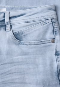 Poche d'un jean en denim bleu clair avec passant de ceinture, double couture et détail de rivet en métal près du bord de la poche.