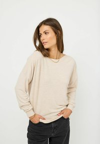 Beige oversized tröja med långa ärmar, ribbade muddar och en enkel halsringning, ihop med mörka jeans. Slät textur, avslappnad design.