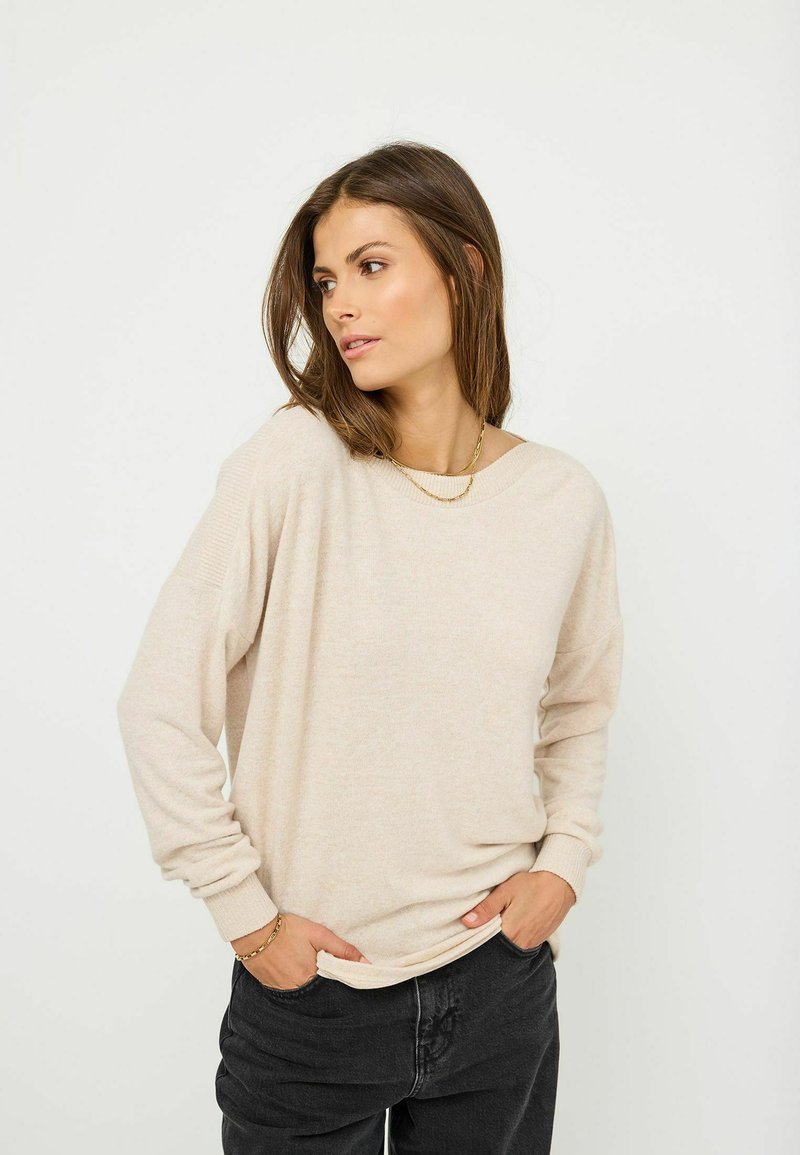 Beige oversized tröja med långa ärmar, ribbade muddar och en enkel halsringning, ihop med mörka jeans. Slät textur, avslappnad design.