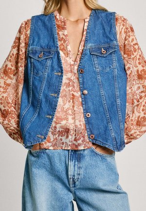 Kvinde iført en blå denimvest over en langærmet bluse med blomsterprint og lyseblå jeans med hænderne i lommerne.