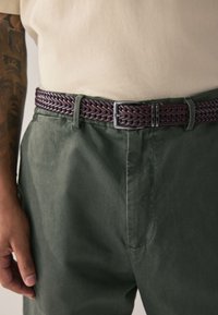 Ceinture en cuir tressé de couleur violet foncé avec une boucle en métal argenté, portée sur un pantalon en coton vert. La ceinture présente un design texturé et une construction robuste.
