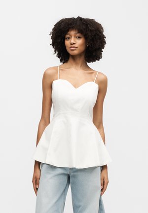 Γυναίκα με κατσαρά μαλλιά που φοράει λευκό αμάνικο τοπ με πεπλούμ και ανοιχτό μπλε τζιν, στέκεται μπροστά σε απλό φόντο.