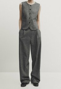 Harmaa ilmanhihaista liiviä, jossa on viisi mustaa nappia, yhdistettynä leveälahkeisiin harmaisiin housuihin. Teksturoitu kangas, jossa on muotoiltu istuvuus ja sivutaskut.