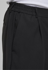 Svarte bukser med elastisk midjebånd, med folder og sidelommer. Laget av glatt, lett stoff med matt finish.