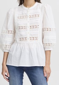 Blusa bianca con un collo alto, maniche a sbuffo, chiusura frontale con bottoni e dettagli in pizzo all'uncinetto sul corpino e sulle maniche. Materiale in cotone morbido.