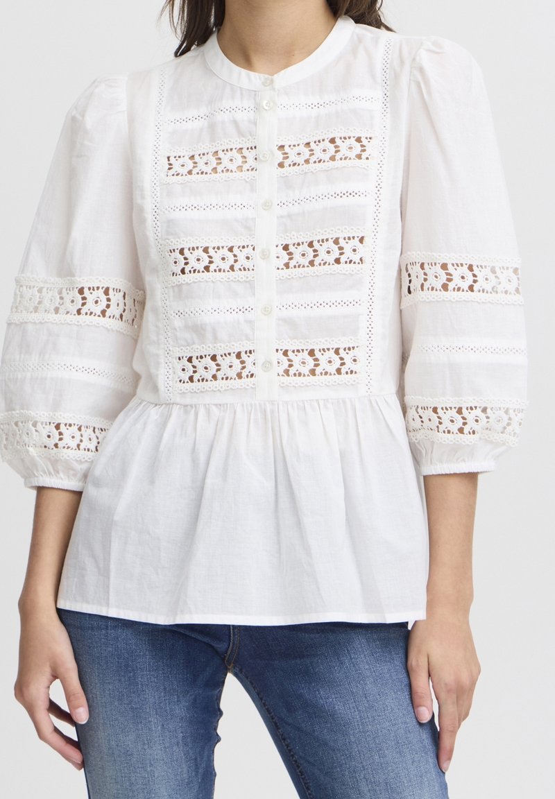 Blusa bianca con un collo alto, maniche a sbuffo, chiusura frontale con bottoni e dettagli in pizzo all'uncinetto sul corpino e sulle maniche. Materiale in cotone morbido.