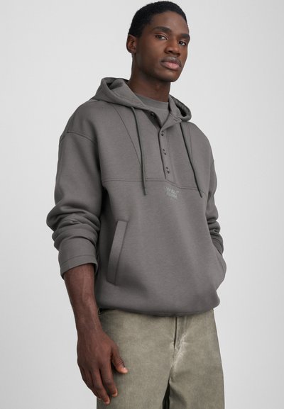 Grijze hoodie met een voorkantzak, knoopsluiting en trekkoorden. Zachte textuur, oversized pasvorm en een klein logo detail op de borst.