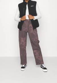 Persona che indossa un gilet in pile nero con chiusura a zip, una maglia bianca a maniche lunghe, pantaloni larghi a fantasia tie-dye viola e scarpe da ginnastica bianco e nero, in piedi su uno sfondo semplice.