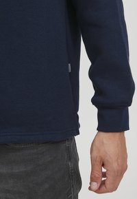 Marineblaues Langarmshirt aus Baumwolle mit rundem Ausschnitt und seitlichen Schlitzen. Der Saum hat eine genähte Kante und ein kleines Etikett an der Naht.
